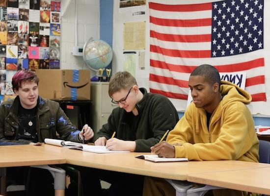 Three 学生 sitting at a table in a classroom, with an American flag in the background. One student in a yellow hoodie is focused on writing, the middle student in glasses is also writing, and the third student with dyed hair is looking up.